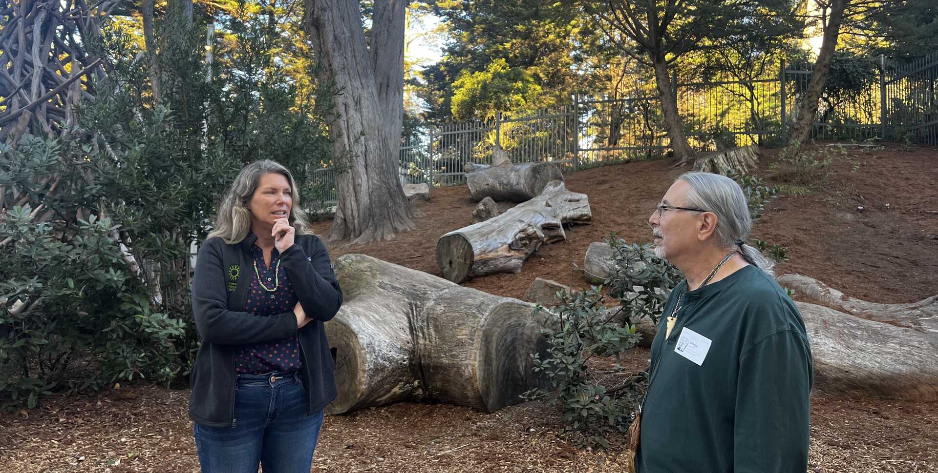 Shannon Tushingham and Gregg Castro are in discussion at Wander Woods, outside the academy in front of tree logs.