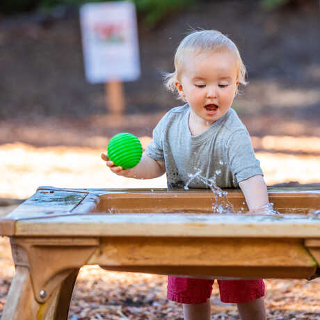 A toddler at play splashes at an outdoor water table in the East Garden