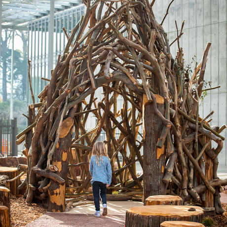 Girl entering fanciful "nest" structure made of woven tree branches in Wander Woods at Cal Academy