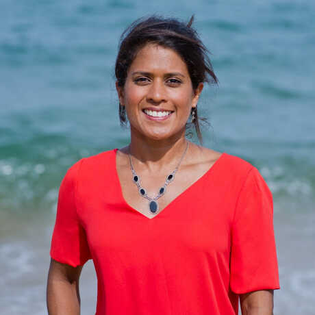 Osher Fellow Dr. Asha de Vos stands on a beach in a read short-sleeved shirt
