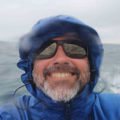 A smiling, bearded Chancey Macdonald wearing sunglasses and a blue hooded rain jacket in front of a large body of water