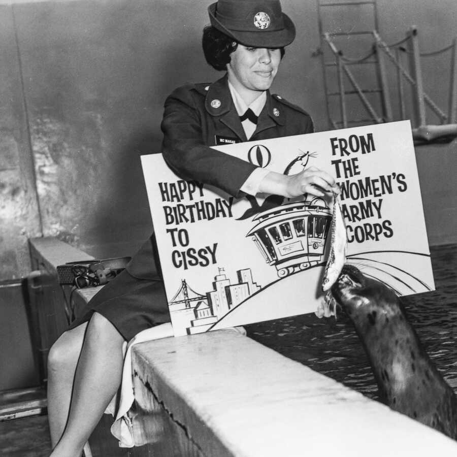 Black and white photo from the 1940s of a woman in uniform feeding fish to a sea lion at the Academy