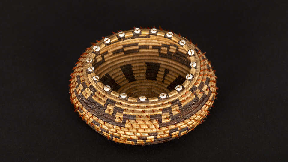 A brown woven Pomo basket is pictured against a black backdrop. This one has little white shells and black stripe-like patterns.