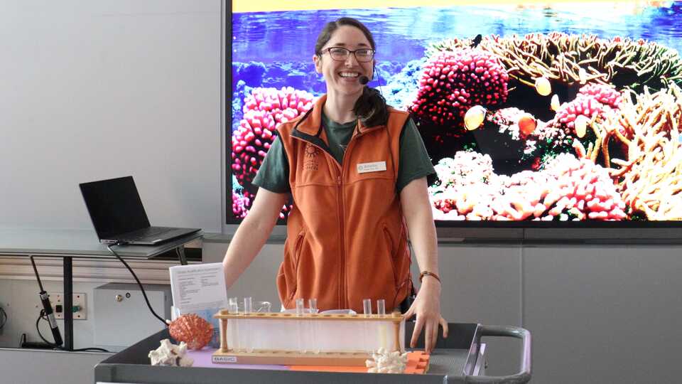 Presenter in orange vest sanding with chemistry set in front and picture of coral behind