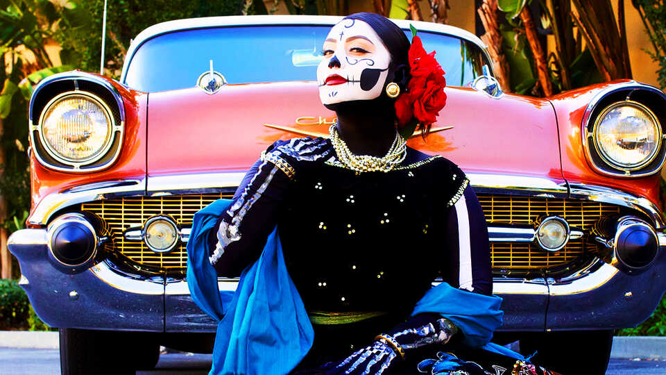 Woman with calavera face paint in traditional dress sitting on ground in front of old red Chevy